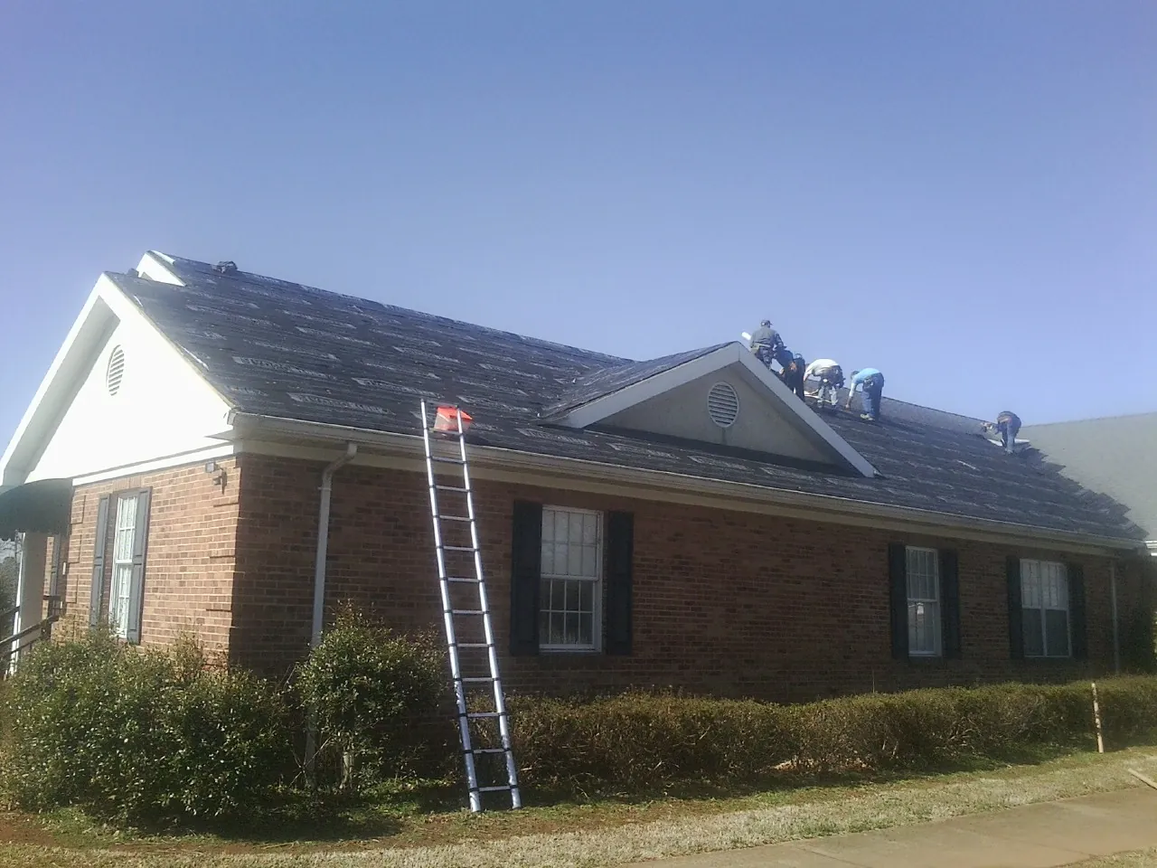 Crew tearing shingles off a brick ranch roof with a ladder set — Metal Roof Repair work by our crew in Friend
