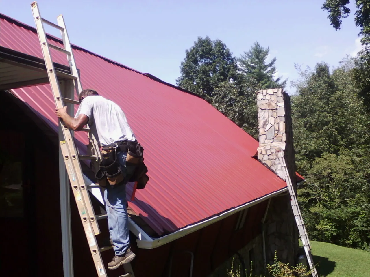Roofer on a ladder beside a red metal roof and stone chimney — Soffit & Fascia Repair by Roofing Brothers in Friend
